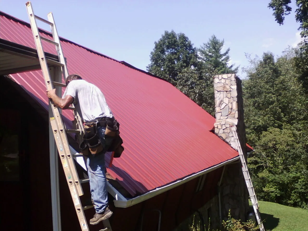 Roofer on a ladder beside a red metal roof and stone chimney — Soffit & Fascia Repair by Roofing Brothers in Long Eddy
